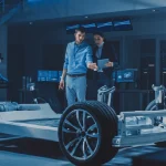 Two automotive engineers stand in front of a vehicle chassis prototype, discussing mobility concepts and vehicle prototypes. The image reflects a scenario within vehicle development and the exploration of new mobility concepts.