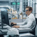 Focused engineer in a white shirt and glasses working on a computer in an industrial setting, possibly analyzing data related to vehicle aerodynamics, with robotic arms and manufacturing equipment visible in the background, indicative of a high-tech automotive development facility.