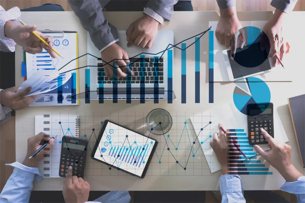Mehrere Personen arbeiten an einem Tisch, auf dem Laptops, Tablets und Dokumente mit Diagrammen liegen. Der Prozess der PSS-Validierung wird durchgeführt, wobei der Fokus auf der Überprüfung von Daten, Analysen und der Potenzialanalyse liegt. Several people are working at a table with laptops, tablets, and documents displaying charts. The image illustrates the PSS validation process, focusing on data, analysis verification, and potential analysis.