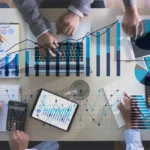 Mehrere Personen arbeiten an einem Tisch, auf dem Laptops, Tablets und Dokumente mit Diagrammen liegen. Der Prozess der PSS-Validierung wird durchgeführt, wobei der Fokus auf der Überprüfung von Daten, Analysen und der Potenzialanalyse liegt. Several people are working at a table with laptops, tablets, and documents displaying charts. The image illustrates the PSS validation process, focusing on data, analysis verification, and potential analysis.