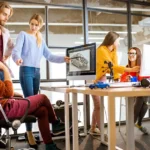 Ein Team von fünf jungen Menschen arbeitet zusammen in einem modernen Büro. Sie diskutieren ein 3D-Design auf einem Monitor, während eine Person eine Visualisierung zeigt. Diese Szene repräsentiert die Beratung in der Konzeptphase, unterstützt durch erweiterte Entscheidungsfindung, um fundierte Projektstrategien zu entwickeln. A team of five young people collaborates in a modern office. They are discussing a 3D design displayed on a monitor while one person showcases a visualization. This scene represents consultation in the concept phase, supported by advanced decision-making to develop well-informed project strategies.