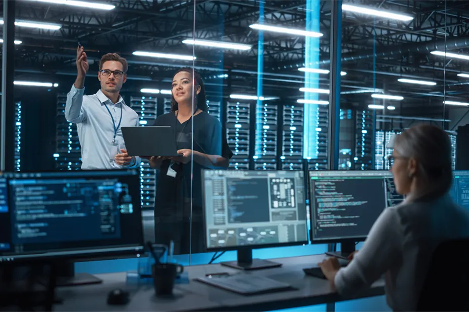 n einem modernen Datenzentrum besprechen zwei Mitarbeitende, einer mit Laptop, die Realisierungsplanung und den Organisation- sowie Infrastrukturplan. Im Vordergrund arbeitet eine weitere Person an einem Computer mit mehreren Bildschirmen, die technische Daten und Systeminformationen anzeigen. In a modern data center, two employees, one holding a laptop, discuss the implementation planning and the organization and infrastructure plan. In the foreground, another person is working at a computer with multiple screens displaying technical data and system information.