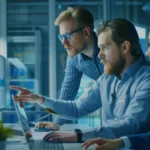 Drei Personen analysieren Diagramme und Daten auf Monitoren, um die PSS-Definition (Product-Service-System) zu erstellen. Dabei führen sie eine Marktanalyse durch und entwickeln Markt-Personas, um das System gezielt auf die Bedürfnisse der Zielgruppen auszurichten. Three people analyze charts and data on monitors to define the PSS (Product-Service-System). They conduct a market analysis and develop market personas to tailor the system to the needs of the target audience.