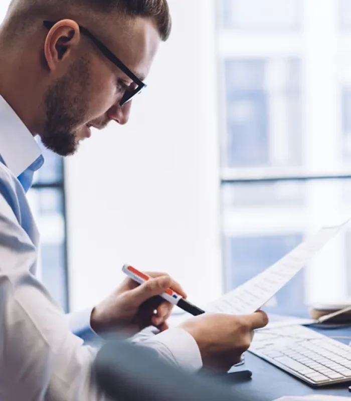 Ein Mann in einem modernen Büro sitzt vor einem Computer und überprüft konzentriert ein Dokument. Auf dem Schreibtisch liegen weitere Papiere und Büromaterialien, während der Prozess der PSS Verification durchgeführt wird. A man in a modern office sits in front of a computer, carefully reviewing a document. Additional papers and office supplies are spread on the desk, as the process of PSS Verification is being performed.