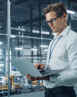 A professional engineer wearing glasses and a white shirt stands in an industrial setting, focused on a laptop. In the background, the manufacturing floor of a vehicle safety testing facility can be seen, indicating the engineer may be analyzing data related to automotive safety performance. Ein professioneller Ingenieur mit Brille und weißem Hemd steht in einer industriellen Umgebung und konzentriert sich auf einen Laptop. Im Hintergrund ist die Produktionshalle einer Prüfeinrichtung für Fahrzeugsicherheit zu sehen, was darauf hindeutet, dass der Ingenieur möglicherweise Daten zur Sicherheit von Kraftfahrzeugen analysiert.